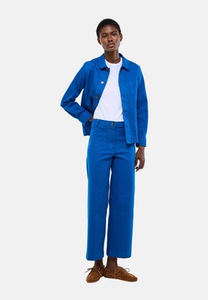 Femme portant une veste bleue à boutons, un pantalon bleu assorti, un T-shirt blanc et des chaussures marron à lacets, debout avec une main dans la poche.