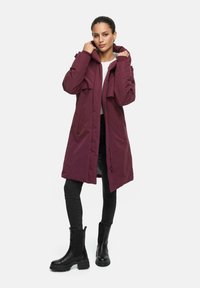 Maroon long coat with a hood, featuring a front zip closure and two large pockets; paired with black skinny jeans and ankle boots.