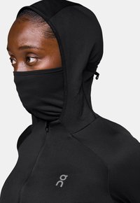 Schwarze Sportjacke mit hohem Kragen, Kapuze und integriertem Gesichtsschutz. Verfügt über einen Reißverschluss und ein dezentes Logo. Glatte Textur.
