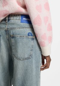 Jasnoniebieskie jeansy z denimu z naszywką z logo HUGO, posiadające tylną kieszeń z niebieskim okrągłym akcentem oraz ręką umieszczoną na pasku w talii.