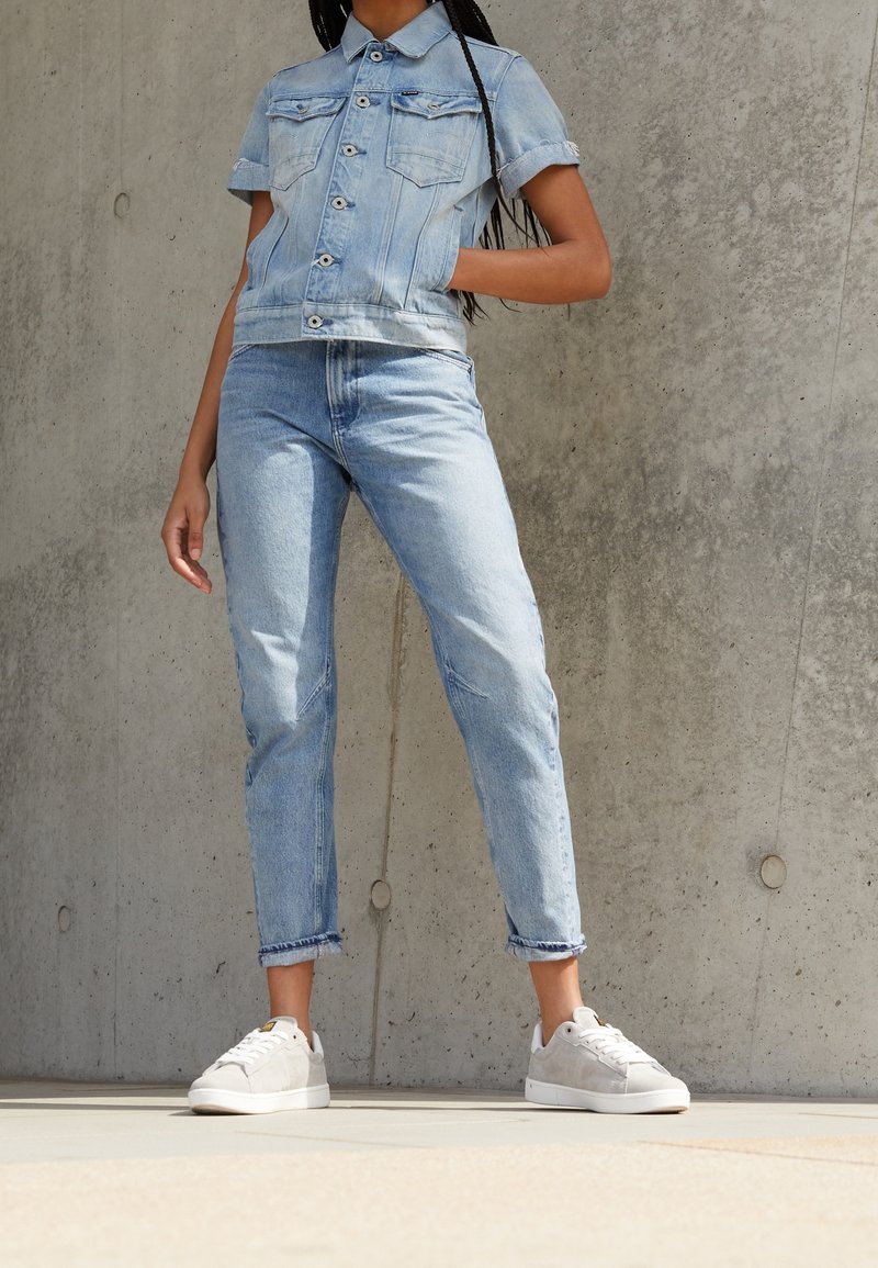 Ljust blå denimjacka med korta ärmar, framknäppning och två bröstfickor, tillsammans med matchande ljusblå jeans och grå sneakers.