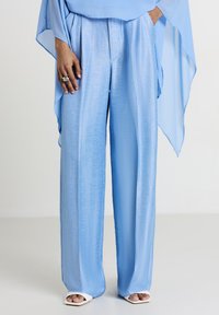 Pantaloni ampi blu chiaro in un tessuto liscio e lucido, con vita alta e fronte plissettata. Indossati con una blusa blu fluida.