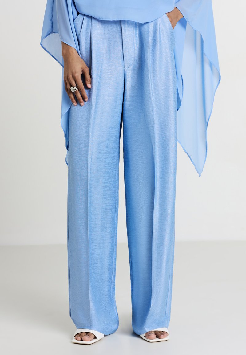 Pantaloni ampi blu chiaro in un tessuto liscio e lucido, con vita alta e fronte plissettata. Indossati con una blusa blu fluida.