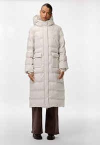 Šviesiai smėlio spalvos pūkinė striukė, iki kelių, su gobtuvu, turinti dideles priekines kišenes ir švarką siuvinėjimu, derinama su tamsiai rudos spalvos kelnių.