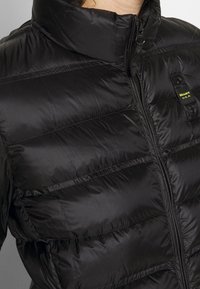 Sort pufferjakke med høj krave, quiltet design, lynlås og logo patch på venstre bryst. Glat, blank overflade.