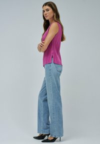 Mujer con cabello largo que lleva una blusa sin mangas de punto rosa, jeans de pierna ancha de color azul claro, tacones negros puntiagudos, de pie de perfil con los brazos cruzados.