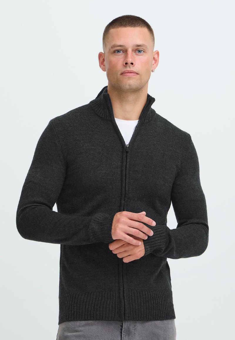 Man met kort haar, gekleed in een donkergrijze ritstrui en lichtgrijze broek, staande met de handen in elkaar gevouwen tegen een effen witte achtergrond.