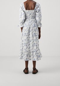 Blommig midi-klänning i vitt med blå blomtryck, smockad rygg, puffärmar och volangkant. Bärs med svarta sandaler.