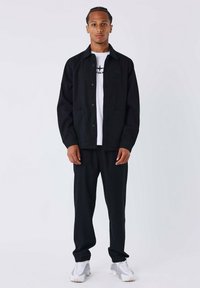 Zwarte jas met knopen, twee voorkeurszakken over een witte t-shirt met een logo, gecombineerd met zwarte broek en grijze sportschoenen.