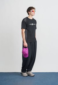 T-shirt desportivo preta com detalhes cinzentos, calças pretas, ténis cinzentos e um boné rosa na mão. Modelo a usar óculos de sol coloridos.