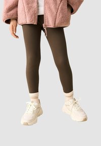 Leggings marroni a coste abbinati a una giacca in pile rosa chiaro e scarpe da ginnastica bianche con dettagli beige. L'outfit presenta calze a righe chiare.