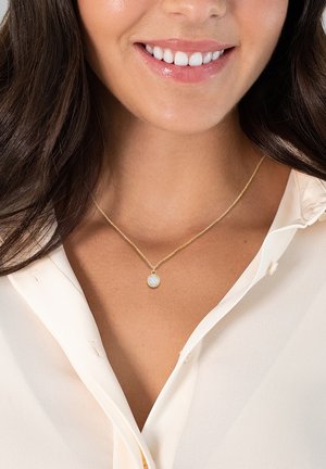 Zlati necklace z okroglim obeskom, ki vsebuje svetlo modri kamen, nosi jo na svetli, nezapeti bluzici. Veriga se zdi nežna in tanka.