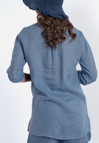 Camicia di lino blu a maniche lunghe, tessuto leggero, orlo dritto e maniche arrotolabili; indossata con un cappello intrecciato blu navy.