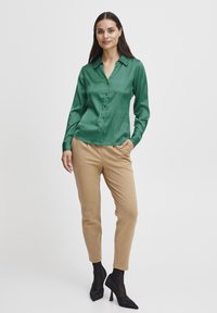 Grünes Satin-Hemd mit langen Ärmeln und Knopfverschluss, kombiniert mit beigen, schmalen Hosen und schwarzen High Heels, vor einem weißen Hintergrund.