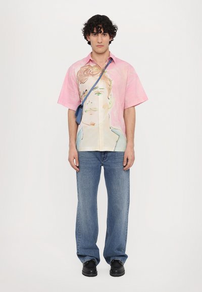 Camisa de manga corta colorida con un degradado en rosa, que presenta un diseño de cara pintada. Combinada con jeans azules y zapatos negros. Incluye un bolso cruzado.
