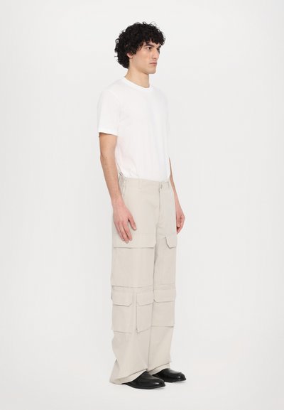 Pantaloni cargo bej cu multiple buzunare, fabricați dintr-un material ușor. Purtați cu un tricou alb și pantofi negri, modelul stă de profil.