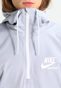 Lichtgrijze Nike sportjack met een hoge kraag, halve rits, verstelbare koorden en een wit logo op de voorkant gedrukt. Soepele textuur.