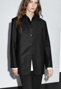 Black wool blend jacket with a collared neck, front buttons, two pockets, and a relaxed fit, paired with a white shirt underneath.