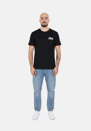 Uomo con testa rasata e barba che indossa una t-shirt nera con grafica, jeans azzurri e sneakers bianche, in piedi rivolto in avanti.