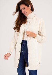 Crème gebreide cardigan met een zachte textuur, knopen aan de voorkant en gebreide manchetten, layered over een witte coltrui, gecombineerd met blauwe spijkerbroek.