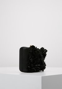 Borsa a pochette rettangolare nera decorata con fiori in tessuto sovrapposti, posizionata su un pedestal bianco contro uno sfondo chiaro uniforme.