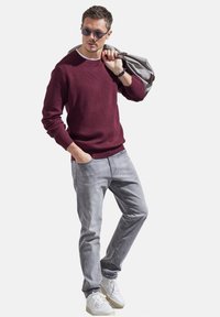 Weinrot strukturiertes Sweatshirt mit rundem Ausschnitt, hellgraue Jeans und weiße Sneakers; hält eine graue Tasche, trägt Sonnenbrillen.