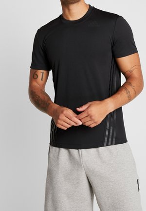 Mann trägt ein schwarzes Adidas-T-Shirt mit seitlichen Streifen und graue Shorts, zeigt Tattoos auf beiden Unterarmen und die Zahl "61" auf dem linken Arm.