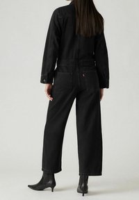 Schwarzer Jeans-Overall mit langen Ärmeln, weitem Bein und Brusttaschen. Verfügt über abgestimmte Nähte und einen entspannten Schnitt. Getragen mit schwarzen Absatzstiefeln.