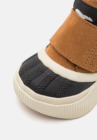 Brun mocka högsko med en svart texturerad gummikappa vid tån, vit mellansula och kardborreband. Distinkta geometriska mönster på tån.