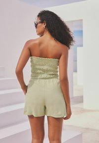Hellgrüner, trägerloser Jumpsuit mit gerafftem Material und gewelltem Saum. Das Model steht vor einem weißen Hintergrund und trägt eine Sonnenbrille.