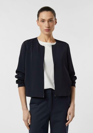 Frau trägt eine schwarze Reißverschlussjacke über einem weißen Hemd mit passenden schwarzen Hosen und steht vor einem schlichten hellen Hintergrund.