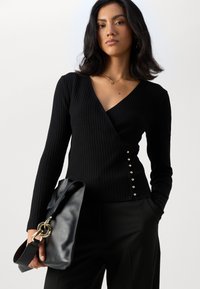 Haut noir à rayures, à manches longues, avec un décolleté en V et des accents de boutons sur le côté. Le mannequin tient un sac en cuir noir avec une chaîne dorée.