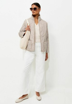 Frau mit übergroßer Sonnenbrille, beiger gesteppter Jacke, weißen weiten Hosen, cremefarbenem Top, trägt große cremefarbene Schultertasche, steht vor weißem Hintergrund.