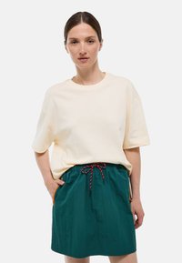 T-shirt oversize crème à manches courtes, associé à une jupe plissée teal avec une poche rehaussée d'orange et une taille à cordon.