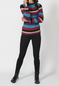 Pull rayé flou avec des bandes rouges, bleues, violettes et noires. Le design est à manches longues et a un col rond. Associé avec un pantalon noir ajusté.
