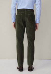 Uomo in pantaloni di velluto a coste color oliva scuro, camicia da donna azzurra chiara infilata dentro, e scarpe eleganti in pelle marrone, in piedi di spalle su uno sfondo neutro.