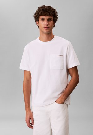 Nooremees lokkis juustega kannab valget lühikeste varrukatega taskuga t-särki ja valgeid pükse, seisab heleda halli tausta ees.