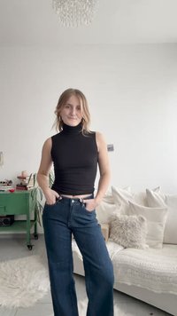 Juoda aptempianti golfas derinamas su tamsiai mėlynais plačiomis kojomis ir aukštu liemeniu džinsais. Kambarys dekoruotas šviesios spalvos pagalvėlėmis ir žaliais baldais.