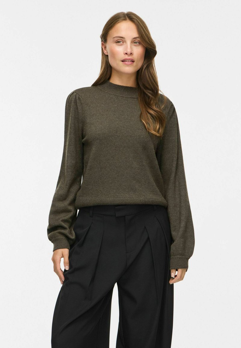 Femme aux longs cheveux, portant un pull vert olive foncé et un pantalon noir large plissé, debout devant un fond clair uni.