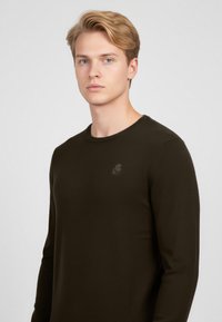 Mladý muž se světlými blond vlasy, oblečený v přiléhavém černém tričku s dlouhým rukávem, stojící před jednobarevným světlým šedým pozadím.