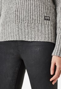Pull gris en tricot avec un ourlet et des poignets côtelés, présentant un petit patch logo noir en bas. Porté avec un jean foncé.