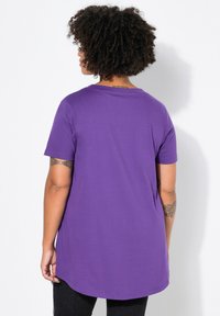 Lakan lilac dengan lengan pendek dan leher bulat, memiliki hem belakang yang lebih panjang, terbuat dari kain halus, dipadukan dengan celana jeans gelap, menampilkan tato di lengan.
