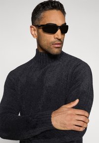 Schwarzer, strukturierter Rollkragenpullover mit gerippten Bündchen und figurnahem Schnitt, kombiniert mit eleganten schwarzen Sonnenbrillen. Modell steht vor neutralem Hintergrund.
