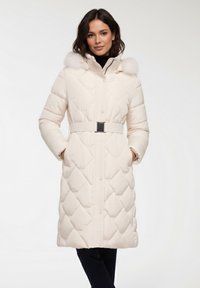 Donna che indossa un cappotto trapuntato color crema con cintura e cappuccio bordato di pelliccia, in piedi con le mani nelle tasche su uno sfondo chiaro e uniforme.