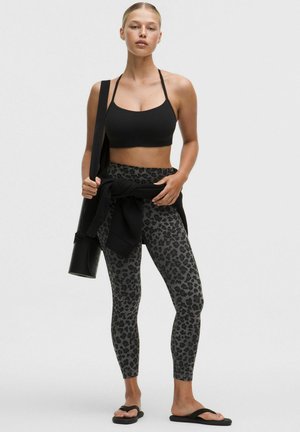 Jeune femme debout portant un soutien-gorge de sport noir, des leggings gris à imprimé léopard, des sandales noires et un sweat-shirt noir noué autour de la taille, tenant un tapis de yoga.