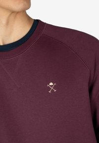Sudadera burdeos con cuello redondo, que cuenta con un interior de color marino texturizado en el cuello. Incluye un pequeño bordado crema de elementos cruzados en el pecho.