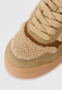 Sneaker beige avec une tige texturée en velours, des accents en cuir lisse, et une semelle en caoutchouc beige clair. Présente des lacets plats et des détails cousus.