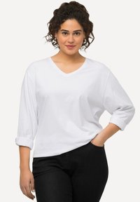 Weißes V-Ausschnitt长期袖上衣 mit umgeschlagenen Manschetten, aus weichem Stoff gefertigt. Kombiniert mit dunklen Jeans, die eine schlanke Passform und Taschendetails aufweisen.