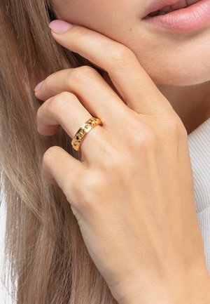 Hand mit goldenem Muster-Ring am Ringfinger nahe den Lippen und langes hellbraunes Haar im Hintergrund.