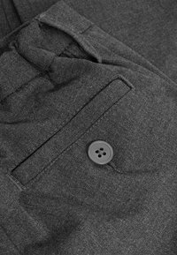 Un pantalon en tissu gris présente une poche avant avec un bouton noir à quatre trous. Le matériau est lisse avec une légère texture et des coutures soignées.